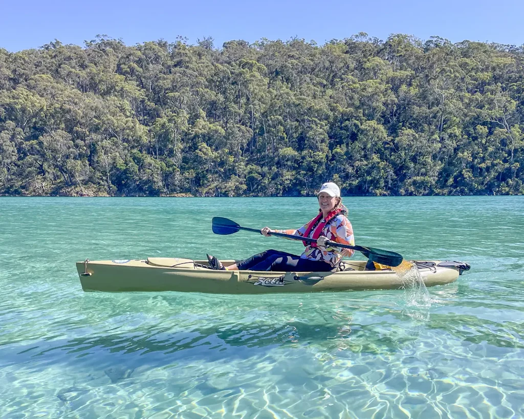 A smiling tour guest paddling a kayak through the crystal-clear, shallow turquoise waters of the Pambula River.