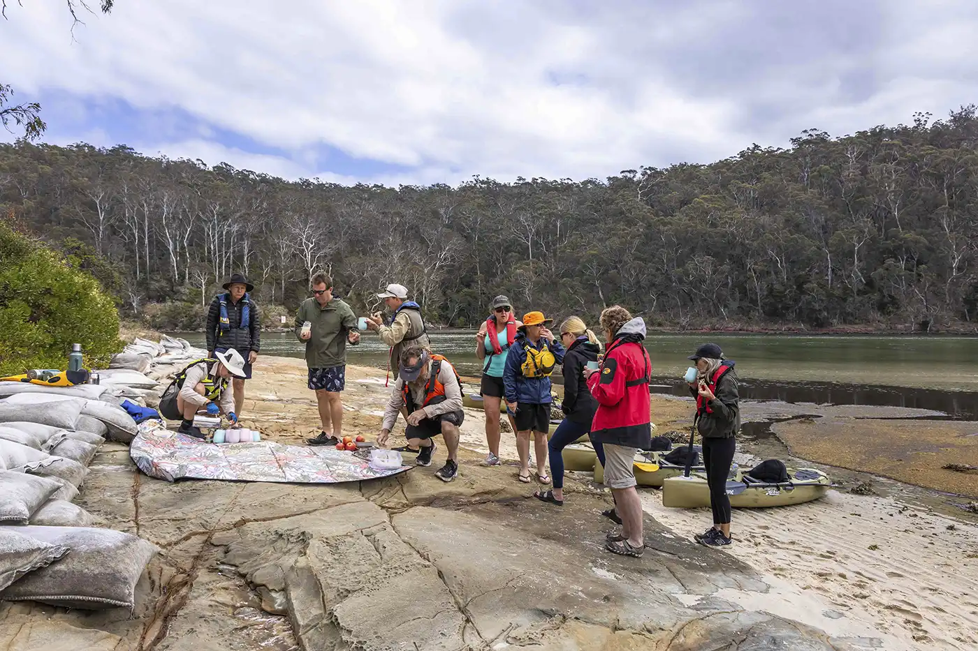 Guests listening attentively to Indigenous guide Nathan Lygon on the beach with Beowa National Park in the background and a picnic rug in the foreground.